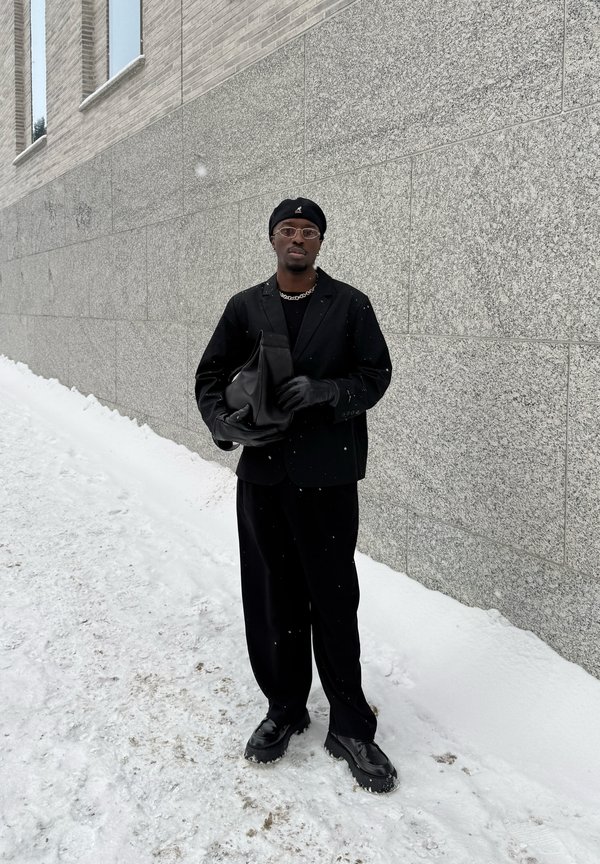 Vyras juodu berete, akiniais, pirštinėmis ir kostiumu, laikantis juodą krepšį, stovintis ant snieguoto žemės šalia pilkos akmeninės sienos.