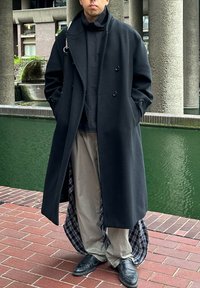 Casaco preto de dupla face, com colarinho grande, combinado com um top preto e calças bege. O forro interior apresenta um padrão xadrez cinza. Calçado preto.