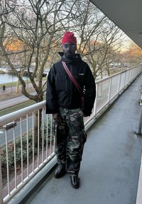 Veste isolante noire avec capuche, pantalon cargo camouflage, bonnet rouge et bottes marron, debout sur un balcon avec des arbres en arrière-plan.