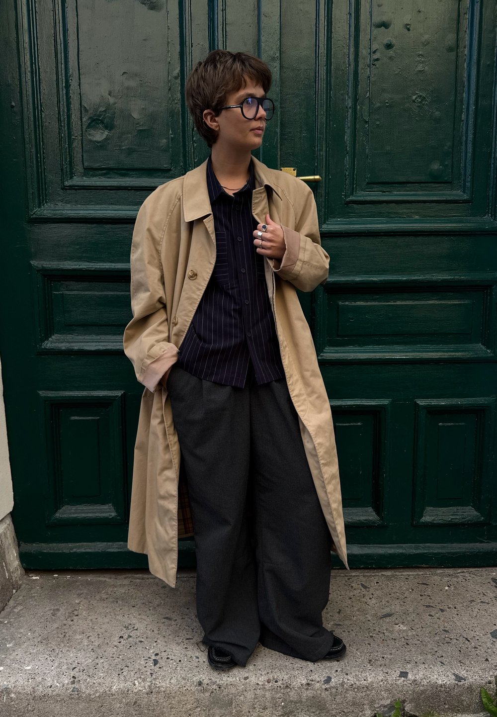 Um casaco trench bege sobre uma camisa preta com riscas, combinado com calças largas escuras e sapatos pretos, em frente a uma porta de madeira verde.