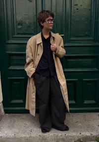 Um casaco trench bege sobre uma camisa preta com riscas, combinado com calças largas escuras e sapatos pretos, em frente a uma porta de madeira verde.