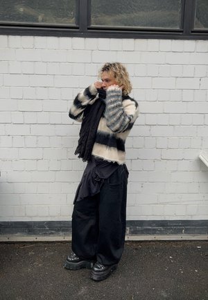 Pull en tricot épais avec des rayures noires et crème, associé à une écharpe noire, un pantalon oversize sombre et des chaussures à plateforme, devant un mur en briques blanches.