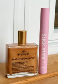 NUXE Huile Prodigieuse Or: klare Flasche, goldene Öl, goldene Akzente. Slick Stick: pinkfarbener zylindrischer Tubus, weiße Schrift, Anti-Flieghaare-Stift, kompaktes Design.