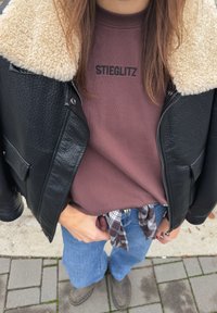 Crna kožna jakna s umjetnim ovratnikom od brtvila, bordo majica s natpisom "STIEGLITZ", plave traperice i sive gležnjače. Karirana košulja vezana oko struka.