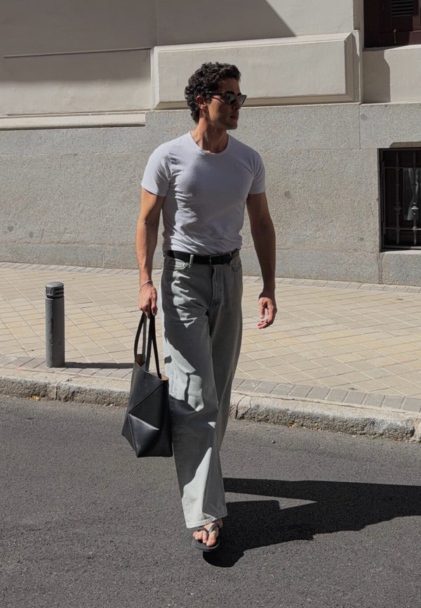 Άνδρας με γυαλιά ηλίου, λευκό t-shirt, φαρδιά ανοιχτόχρωμα τζιν και σανδάλια περπατά στον δρόμο κρατώντας μαύρη τσάντα tote στο ηλιακό φως.