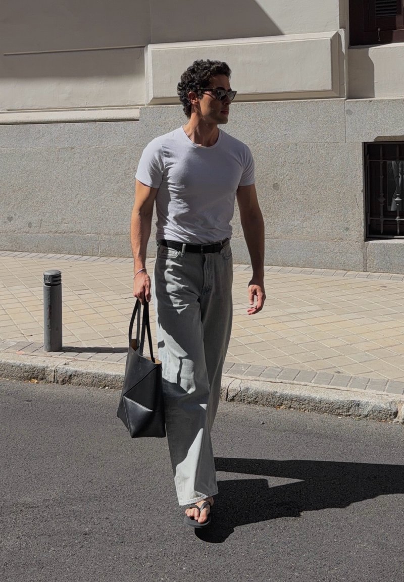 Homme portant des lunettes de soleil, un t-shirt blanc, un jean large clair et des sandales, marche dans la rue en tenant un sac fourre-tout noir au soleil.