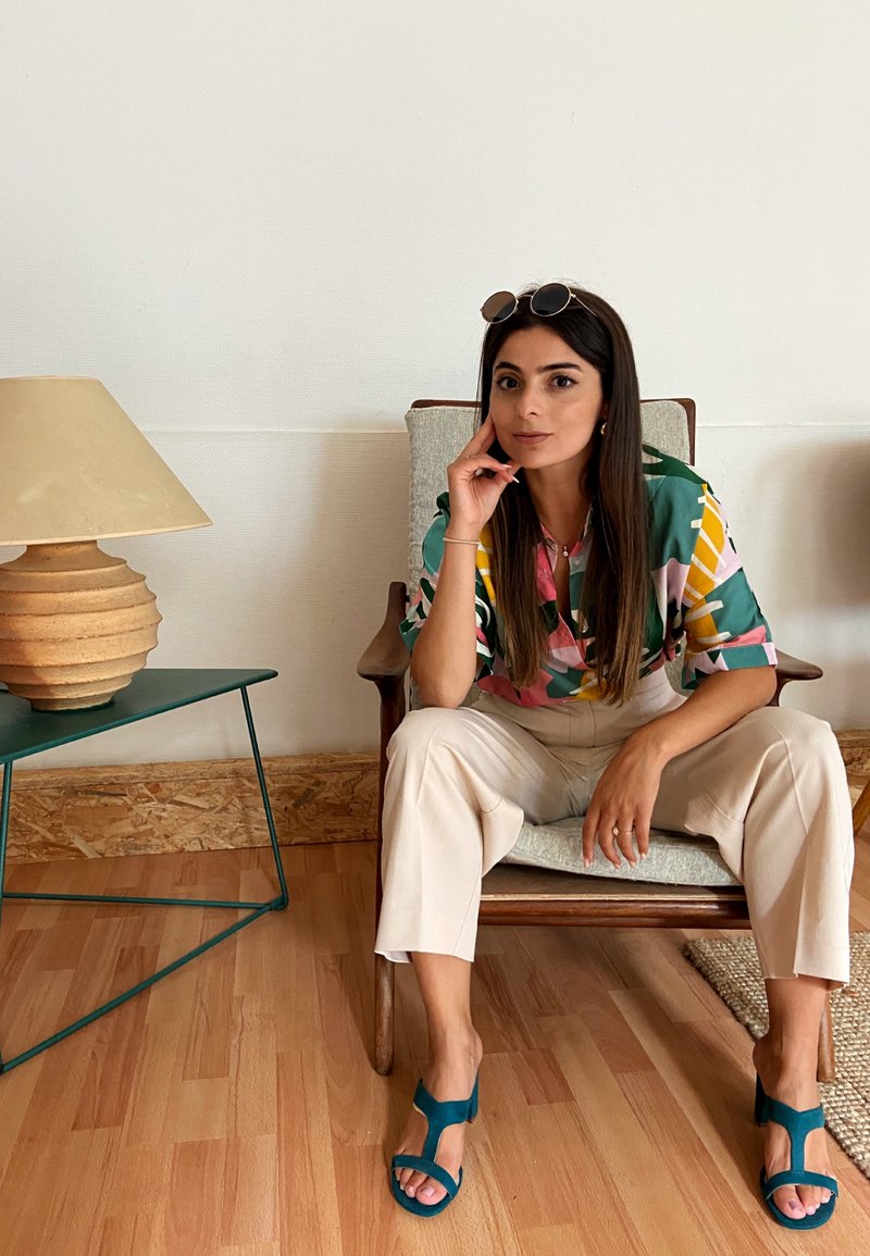 Többszínű, zöld, rózsaszín és sárga mintás ing; krémszínű nadrág; téglakék nyitott orrú cipők. Fa szék, kerek bézs lámpa, zöld éjjeliszekrény.