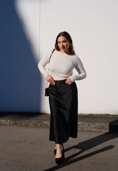Top blanco de manga larga con textura acanalada combinado con una falda maxi de satén negra. Un cinturón dorado acentúa la cintura, complementado con tacones negros y un bolso pequeño.
