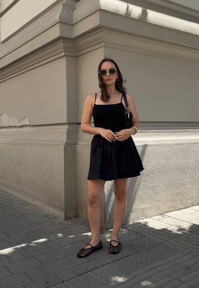 Vestido negro sin mangas con un corpete ajustado y una falda acampanada. Combinado con zapatos negros de malla. Los accesorios incluyen un pequeño bolso negro y gafas de sol.