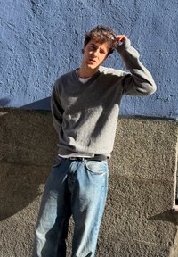 Pull gris à col en V porté sur un t-shirt blanc, assorti à un jean bleu ample. Le tissu semble texturé contre un mur bleu et une surface beige rugueuse.