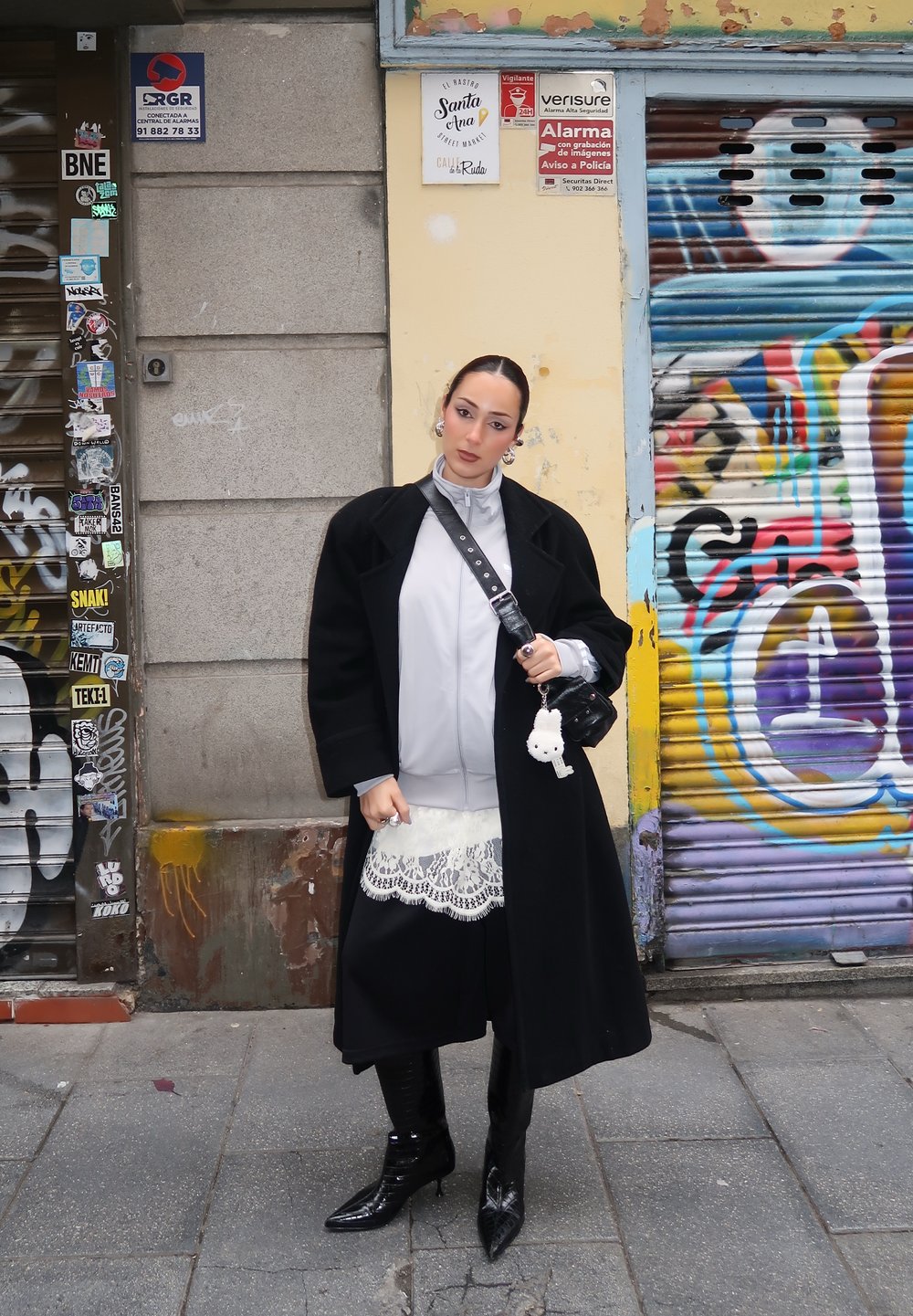 Femme portant un manteau noir, une robe en dentelle blanche, des bottes noires et un sac bandoulière, debout dans une rue urbaine couverte de graffitis.