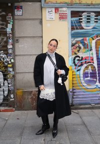 Femme portant un manteau noir, une robe en dentelle blanche, des bottes noires et un sac bandoulière, debout dans une rue urbaine couverte de graffitis.