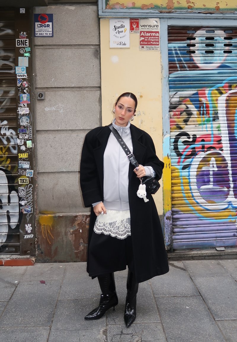 Femme portant un manteau noir, une robe en dentelle blanche, des bottes noires et un sac bandoulière, debout dans une rue urbaine couverte de graffitis.