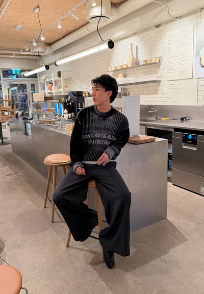 Persona con suéter negro y pantalones anchos sentada en un taburete en la barra de un café moderno con estantes, máquinas de café y un menú en la pared de ladrillo blanco.