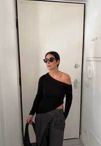 Haut noir à côtes décolleté épaules, pantalon plissé gris et lunettes de soleil noires. Le décor comprend une porte blanche simple et un mur blanc.