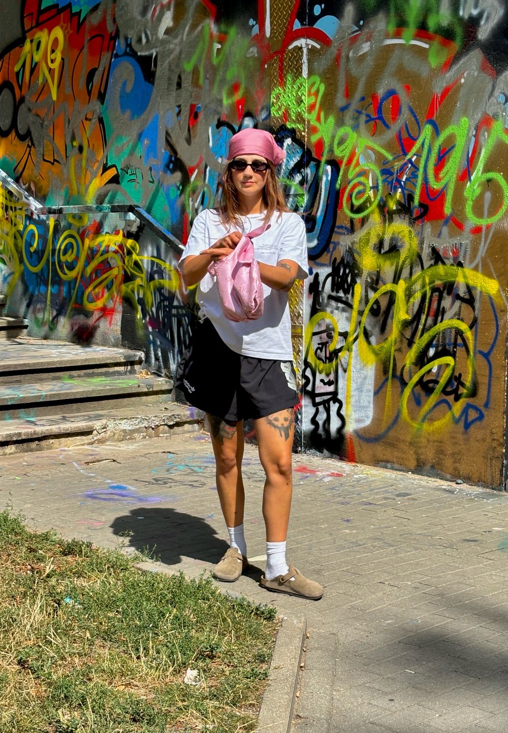 Jednotlivec oblečený v bielom tričku, čiernych šortkách a hnedých topánkach, drží ružovú látku a stojí pred farebnou graffiti stenou.