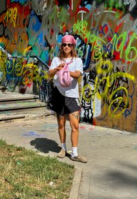Jednotlivec oblečený v bielom tričku, čiernych šortkách a hnedých topánkach, drží ružovú látku a stojí pred farebnou graffiti stenou.