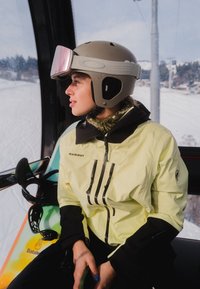 Personne portant une veste de ski jaune et un casque avec des lunettes de ski reposant sur la tête, assise à l'intérieur d'une cabine de télésiège tenant un snowboard.