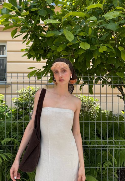 Vestido blanco de lino sin tirantes con un diseño ajustado, combinado con un bolso de hombro de ante marrón oscuro. Ambientado en un entorno con vegetación y una cerca.