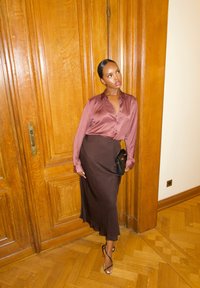 Jedwabna bluzka w kolorze mauve, zestawiona z brązową satynową spódnicą, noszona z czarną kopertówką i sandałkami na obcasie, na tle drewnianej ściany.