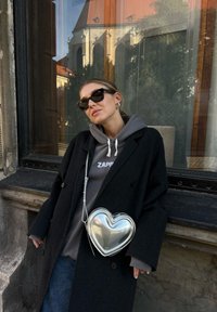 Svart ullrock över en grå hoodie, med en glänsande silverhjärtformad väska. Solglasögon och minimalistiska örhängen, mot en fönsterbakgrund.