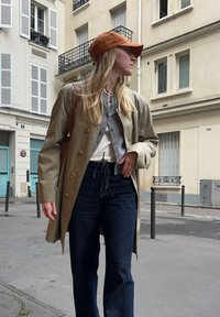 Trench coat beige chiaro con bottoni dorati, cardigan grigio, top bianco, jeans blu scuro e cappellino da baseball marrone. Ambientazione di strada con edifici.