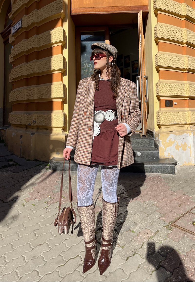 Žena oblečená v kockovanom saku, bordových šatách, širokom striebornom opasku, bielych čipkovaných pančuchách, vzorovaných topánkach, kockovanej baretke a nesie hnedú kabelku vonku.