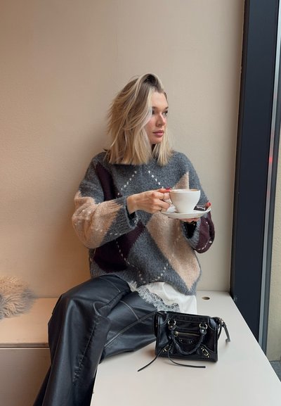 Suéter grueso de punto con patrón de rombos en gris, beige y burdeos. Pantalones de cuero negros, bolso pequeño negro y taza blanca con platillo.