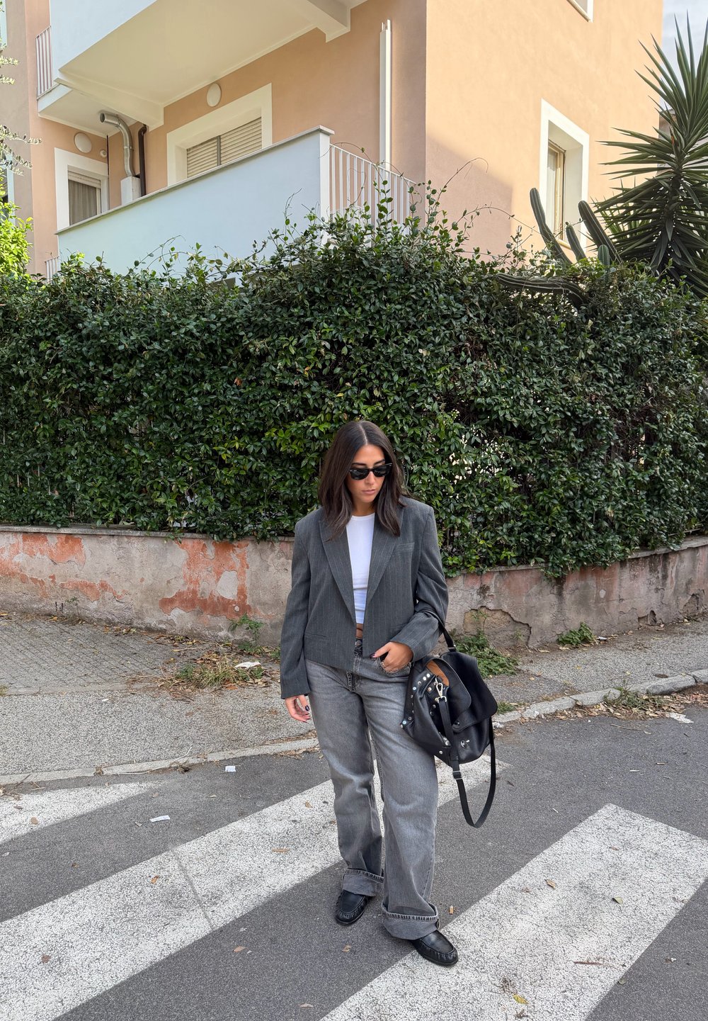 Grijze cropped blazer over een witte top, gecombineerd met grijze wijde jeans en zwarte schoenen. Accessoires omvatten een zwarte handtas en een zonnebril.