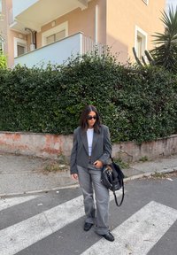 Grijze cropped blazer over een witte top, gecombineerd met grijze wijde jeans en zwarte schoenen. Accessoires omvatten een zwarte handtas en een zonnebril.