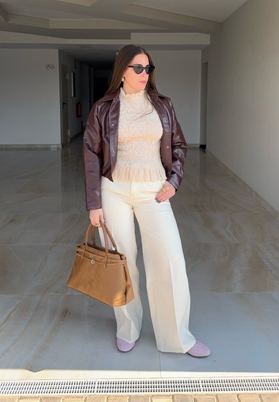 Mujer con chaqueta de cuero marrón, blusa crema con volantes y pantalones, zapatos lila, gafas de sol oscuras, sosteniendo un gran bolso marrón en interiores.