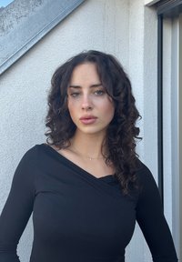 Haut noir à manches longues avec un col drapé, porté avec un délicat collier en or. Des cheveux bouclés encadrent le visage sur un fond de mur texturé.