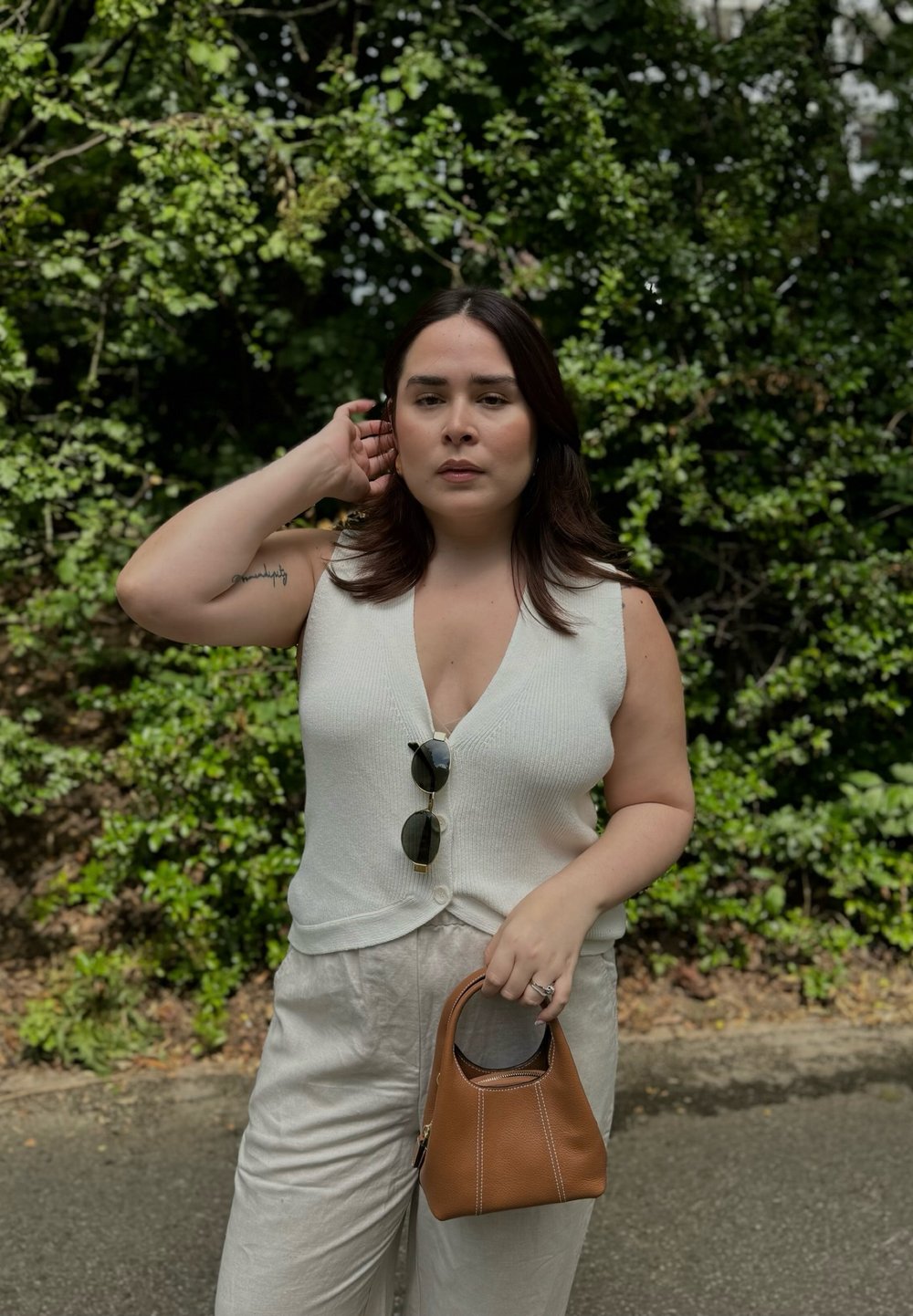 Haut en tricot sans manches blanc avec un décolleté en V et des détails de boutons, associé à un pantalon large de couleur claire. Tient un petit sac à main en cuir marron.