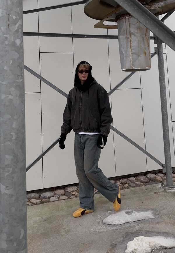 Ung person iført sort hættetrøje, handsker, solbriller, jeans og gule sneakers, stående ved en industriel metalstruktur og grå vægpaneler.
