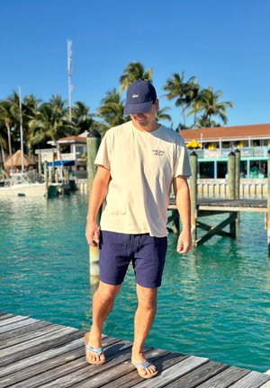 Hombre con pantalones cortos, camiseta y gorra caminando por un muelle de madera junto a aguas turquesas con botes y palmeras al fondo.