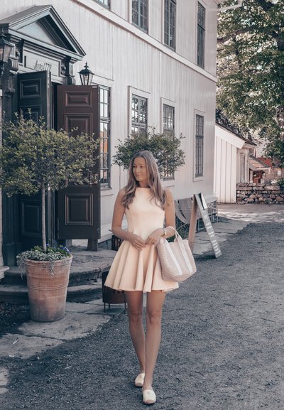 Vestido sin mangas de color rosa con una silueta acampanada, bolsa de mano tejida con acentos blancos, rodeada de vegetación y arquitectura histórica.