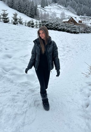 Mujer con abrigo de invierno negro, guantes y botas caminando por un sendero cubierto de nieve, con árboles cubiertos de nieve y una casa de madera al fondo.