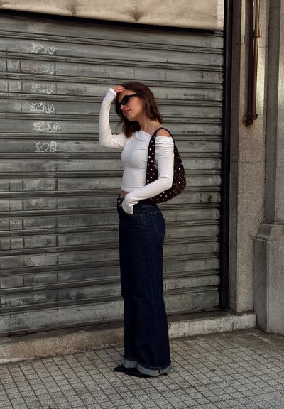 Mujer con blusa blanca de hombros descubiertos, jeans oscuros de pierna ancha y gafas de sol, con una mano en la cabeza, posando frente a una persiana metálica cerrada.