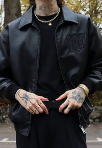 Sort bomberjakke i imiteret læder med lynlåslukning, ribstrikkede manchetter og et broderet logo. Bær over en sort skjorte, som fremviser tatoverede hænder.