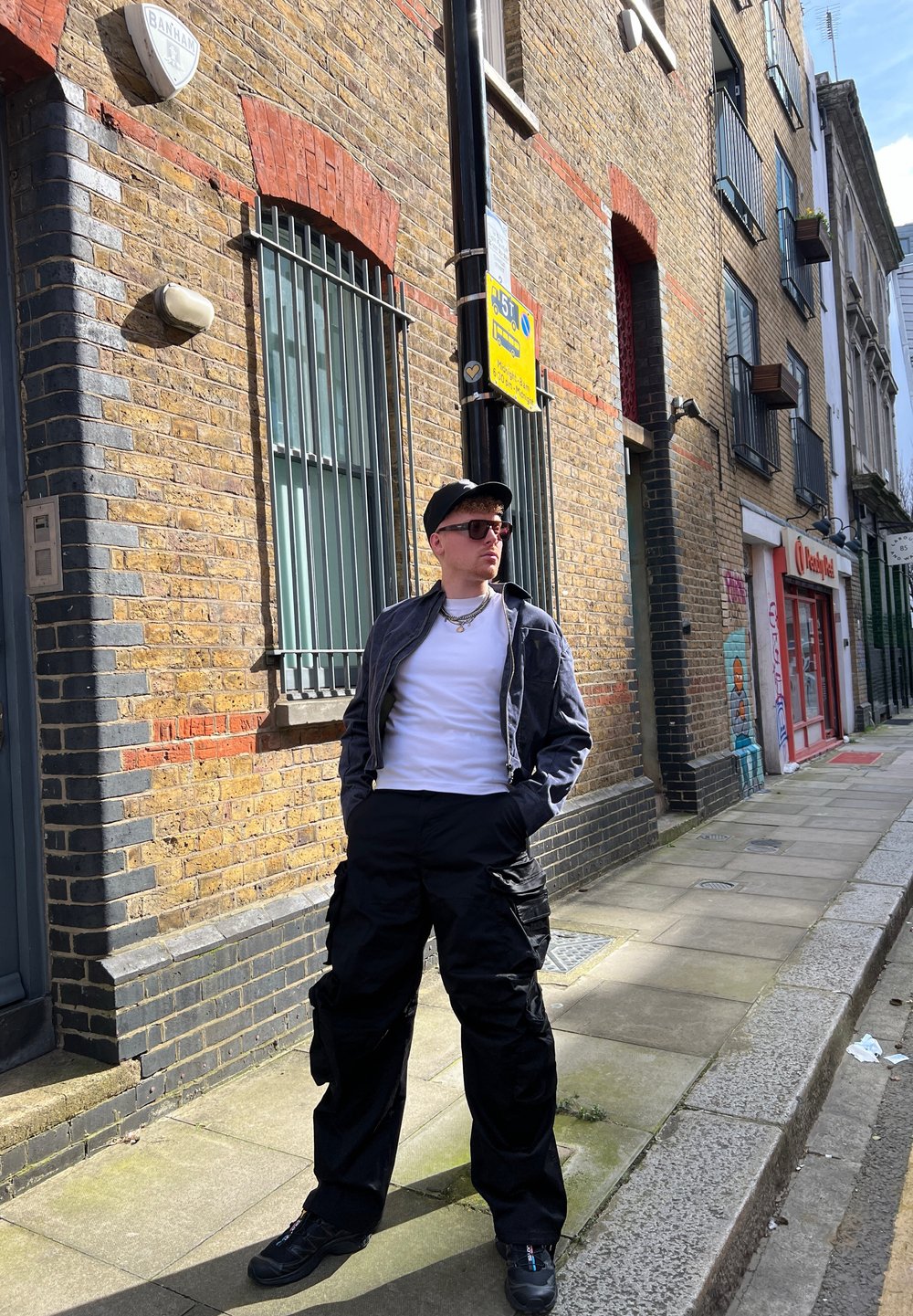 Καθημερινό ντύσιμο που περιλαμβάνει μια γκρι bomber jacket, λευκό t-shirt, μαύρα cargo παντελόνια και αθλητικά παπούτσια, σε φόντο από τούβλινο τοίχο σε έναν αστικό δρόμο.