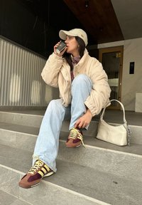 Κρέμ κοτλέ σακάκι, ροζ καρό πουκάμισο, ανοιχτό μπλε τζιν, μπορντώ και μπεζ αθλητικά παπούτσια με κίτρινες κορδόνες, μπεζ τσάντα χειρός, κρατώντας μια κούπα καφέ.