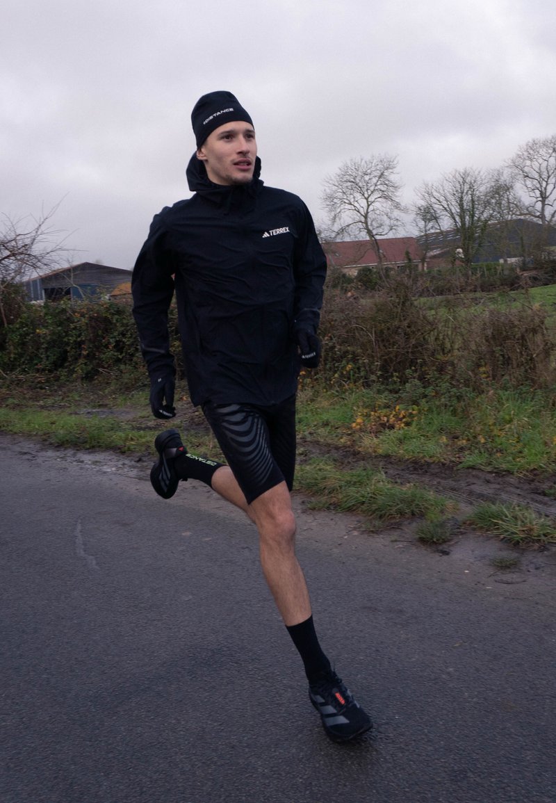 Homme portant une tenue de course noire et des gants, jogging sur une route à côté d'herbe et d'arbres nus sous un ciel couvert.