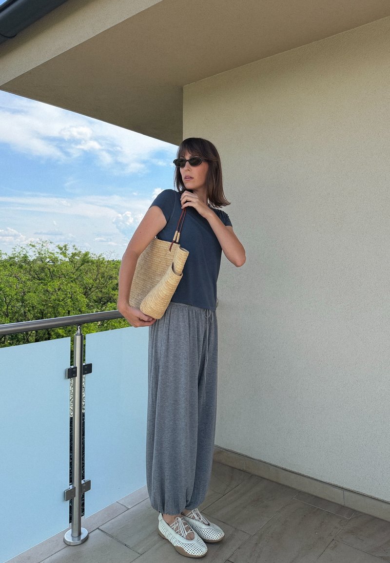 Frau mit dunklem Haar und Sonnenbrille steht auf dem Balkon, trägt einen dunklen Oberteil, graue lockere Hosen, weiße Schuhe und hält eine geflochtene Strohtasche über der Schulter.