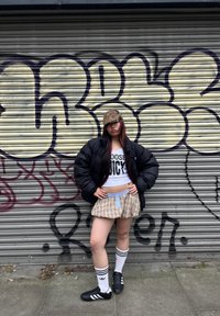 Casaco puffer preto, top curto branco com a inscrição "CHOSE JUICY", saia mini xadrez, meias brancas com riscas pretas e sapatos de desporto pretos.