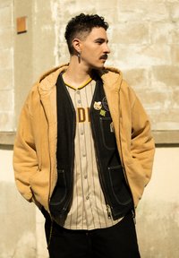Mężczyzna o krótkich, kręconych włosach i wąsach, ubrany w beżowy sweatshirt, czarną kamizelkę z przypinkami oraz w paskowaną koszulę, stoi przy kamiennej ścianie.