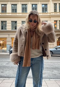 Γυναίκα με γυαλιά ηλίου που φοράει ένα μπεζ γούνινο παλτό, καφέ κασκόλ, λευκό πουλόβερ και τζιν, στέκεται σε αστική οδό με κτίρια στο βάθος.