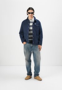 Námornícky modrá bunda s gombíkovou prednou časťou a dvomi vreckami, skombinovaná s prúžkovanou košeľou, svetlomodrými džínsy a hnedými topánkami. Nosené tmavé slnečné okuliare.