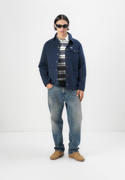 Chaqueta azul marino con cierre de botones y dos bolsillos, combinada con una camisa de rayas, jeans de color azul claro y zapatos de color beige. Se llevan gafas de sol oscuras.