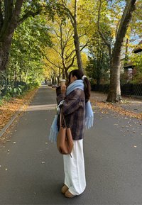 Model v rjavi karo jakni, svetlo modrem šalu, belih širokih hlačah in rjavih škornjih, ki drži skodelico kave, stoji na poti obkroženi z drevesi.