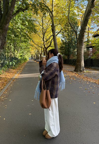 Modelo con una chaqueta de cuadros marrón, bufanda azul claro, pantalones blancos de pierna ancha y botas marrones, sosteniendo una taza de café, de pie en un camino bordeado de árboles.